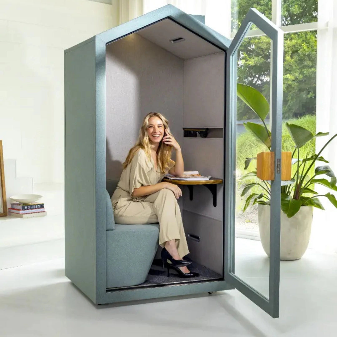 The product is an acoustic meeting pod designed for private conversations or work. It features a cozy interior with sound-absorbing materials, a small desk, and a comfortable seat. The scene shows a woman sitting inside the pod, smiling and talking on the phone, with a plant and some books visible in the background.
