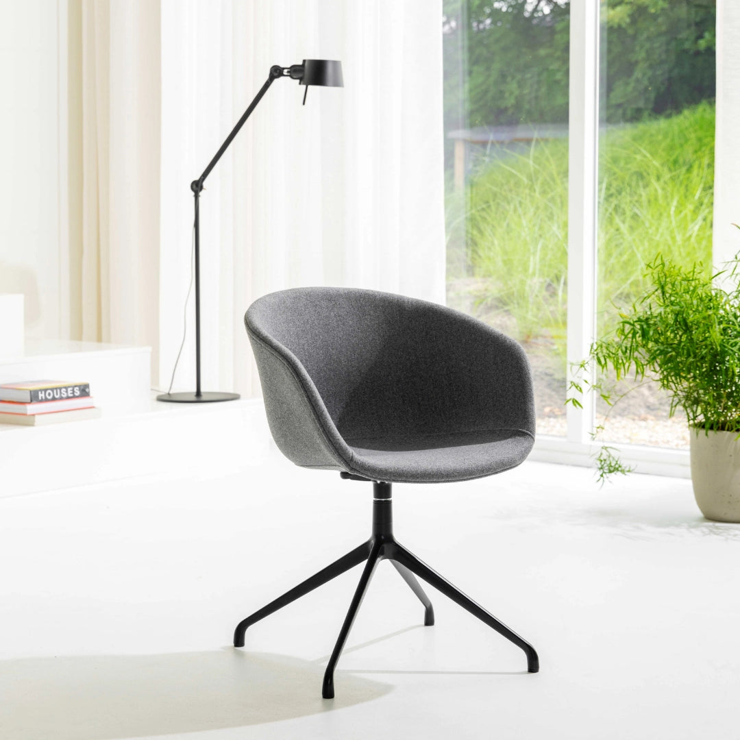 The product is an ergonomic office chair, characterized by its modern design and comfortable, rounded seat. It features a gray fabric upholstery and a sleek black base with a four-legged design. The chair is positioned in a bright space with natural light, accompanied by a standing lamp and some greenery in the background, creating a stylish and inviting atmosphere.