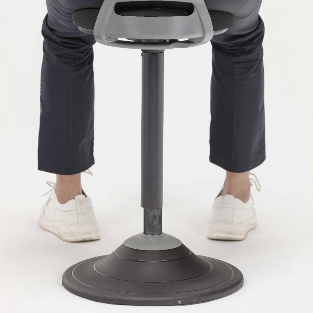 The image depicts a sit-stand stool designed for ergonomic support, promoting active sitting. The stool features a sleek design with a circular base and an adjustable height mechanism. In the image, a person is shown using the stool, wearing dark pants and white shoes, emphasizing the product's use in a modern workspace.