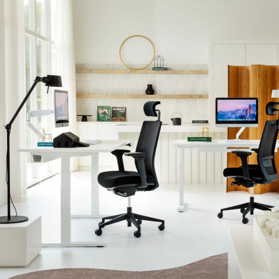 The image features an ergonomic office chair, likely designed for comfort and support during long hours of work. The brand and model are not specified, but the chair is black with a high back and adjustable features. In the background, there are two desks with monitors, a stylish lamp, and decorative items on shelves, creating a modern office environment.