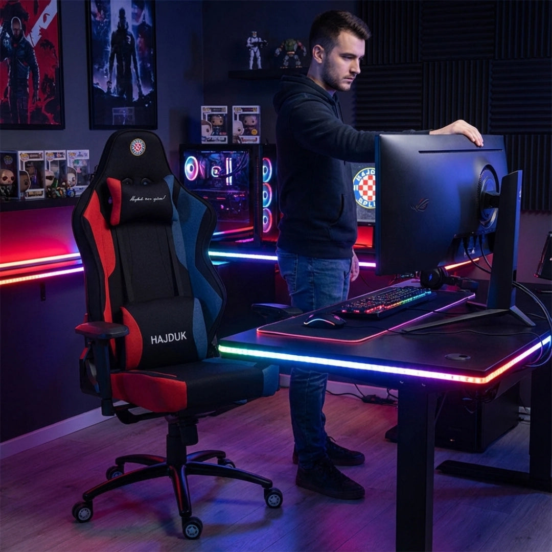 The image features an ergonomic gaming chair from the brand Hajduk, model UVI X. The chair is designed with a combination of red and blue accents and is positioned in a modern gaming setup, complete with LED lighting and a sleek desk. A person is seen adjusting a monitor while the gaming chair is prominently displayed in the foreground.