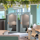 The image features acoustic meeting pods designed for private discussions in office environments. These pods have a cottage-like design with a soft gray exterior and wooden accents. Inside, they are furnished with seating and a small table, providing a cozy and soundproof space for meetings or focused work. The setting includes a modern office with greenery and ergonomic furniture.