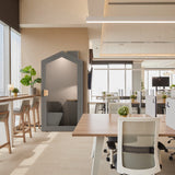 The image features an acoustic meeting pod, specifically the "Cottage" model. This product is designed for private and soundproofed meetings in office environments. The pod has a house-like shape, with a gray exterior and a cozy interior that includes seating and a small desk, providing a quiet space amidst a modern office setting.