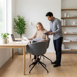 The image features an ergonomic office chair in a modern workspace setting. A woman is seated at a wooden desk, reviewing documents, while a man stands beside her, holding a tablet. The room has a minimalist design with a plant and shelves in the background, creating a bright and inviting atmosphere.