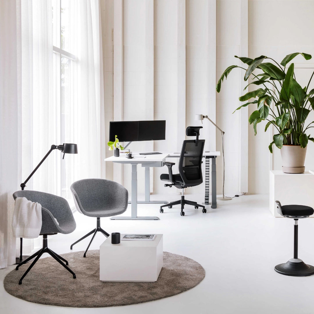 The image features an ergonomic office chair, likely designed for conference or professional settings. The chair has a sleek, modern design with a mesh back for breathability. The workspace includes a standing desk with dual monitors, a stylish lamp, and a cozy seating area with two gray chairs and a white coffee table, all set in a bright, minimalist environment with