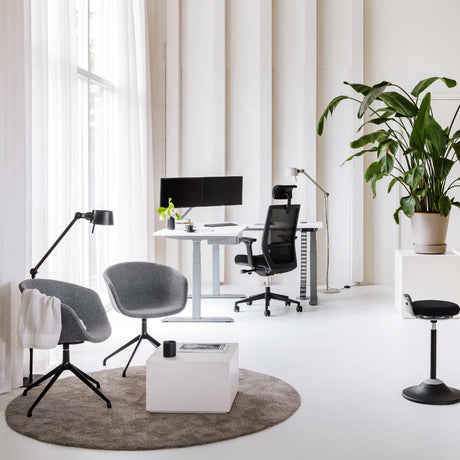 The image features an ergonomic office chair, likely designed for conference or professional settings. The chair has a sleek, modern design with a mesh back for breathability. The workspace includes a standing desk with dual monitors, a stylish lamp, and a cozy seating area with two gray chairs and a white coffee table, all set in a bright, minimalist environment with