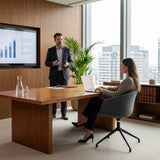 The image features an ergonomic office chair, likely designed for conference or meeting settings. The chair has a modern design with a fabric seat and a five-point base. In the scene, a woman is seated at a wooden conference table, working on a laptop, while a man stands nearby presenting information displayed on a screen. The office environment is sleek and professional