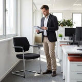 The image features a conference chair with a sleek, modern design, likely intended for office or meeting environments. It has a black fabric seat and backrest supported by a chrome frame. A man in business attire stands nearby, reviewing documents, with a well-lit office setting visible in the background, including desks and plants. The overall atmosphere suggests a professional workspace