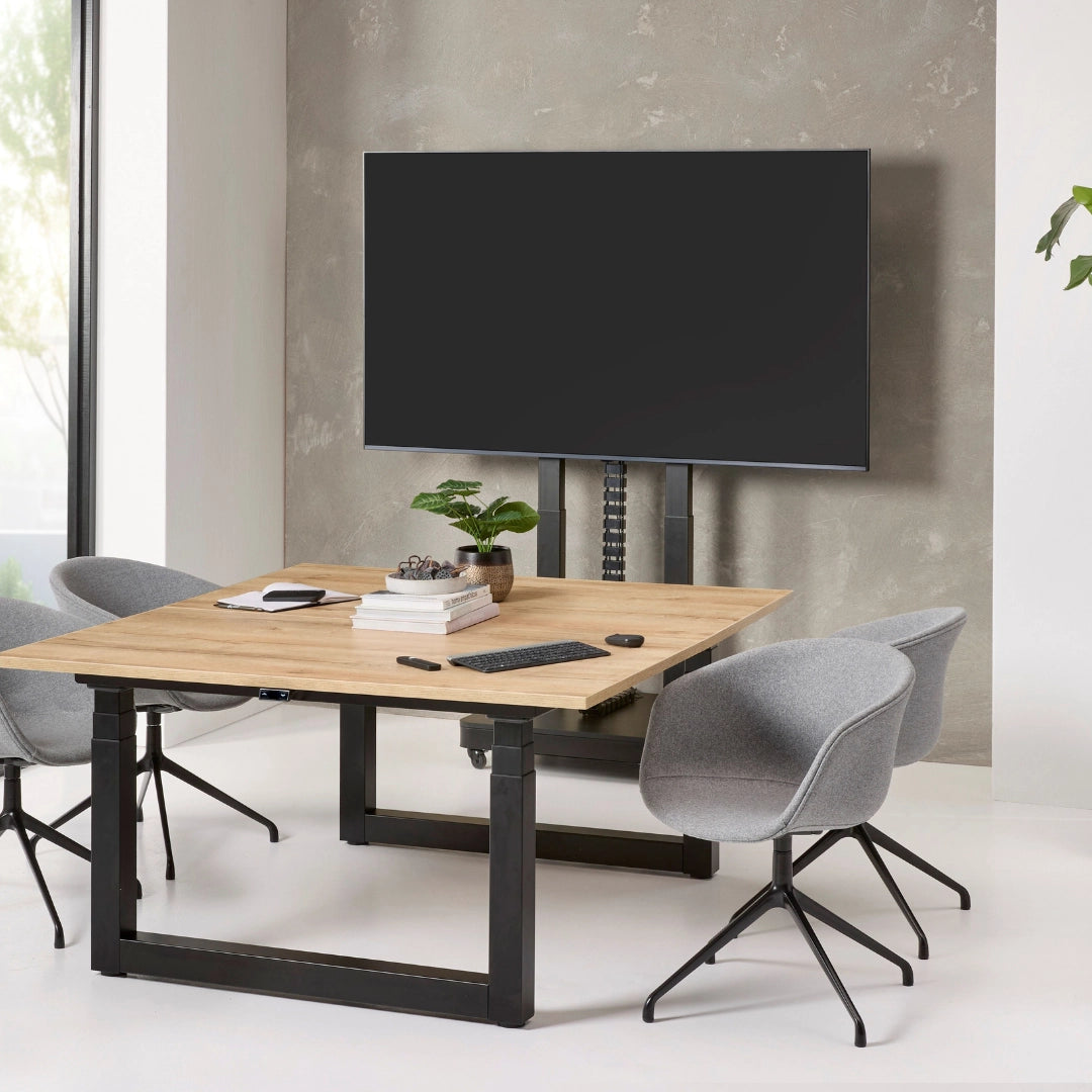 The image features an electric mobile TV lift stand, designed for mounting televisions. It showcases a sleek, modern design with a wooden table and two gray chairs. The TV is positioned above the table, supported by an adjustable lift mechanism, creating a functional and stylish workspace.