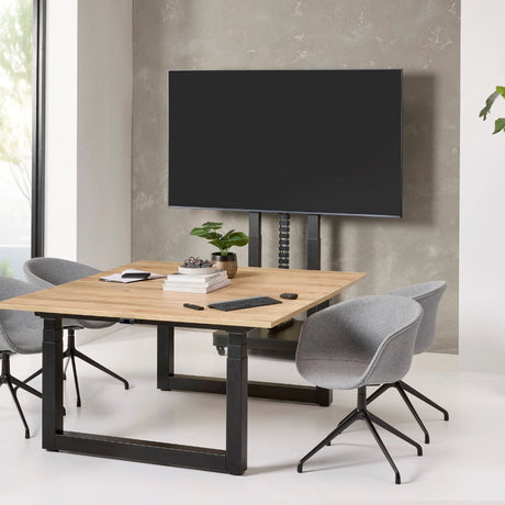 The image features an electric mobile TV lift stand, designed for mounting televisions. It showcases a sleek, modern design with a wooden table and two gray chairs. The TV is positioned above the table, supported by an adjustable lift mechanism, creating a functional and stylish workspace.