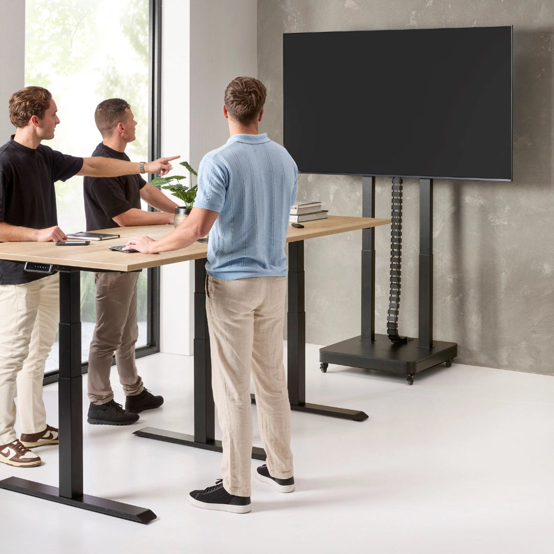 The product is an electric mobile TV lift stand. It features a sleek, modern design with a sturdy base and adjustable height, allowing for easy positioning of the TV. In the image, three individuals are interacting around a standing desk, pointing at the TV mounted on the lift stand, which is currently turned off. The setting appears contemporary, with large windows