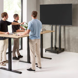 The product is an electric mobile TV lift stand. It features a sleek, modern design with a sturdy base and adjustable height, allowing for easy positioning of the TV. In the image, three individuals are interacting around a standing desk, pointing at the TV mounted on the lift stand, which is currently turned off. The setting appears contemporary, with large windows