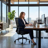 The image features an ergonomic office chair, specifically the "Vane" model. It showcases a woman working at a desk with a laptop and two monitors in a modern office setting, characterized by large windows and a view of the city. The chair is designed for comfort and support, highlighting its ergonomic features.