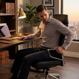 The image features an ergonomic executive office chair, designed for comfort and support during long hours of work. A man is seated in the chair, adjusting it while working at a desk with a laptop. The setting includes a stylish office with a warm ambiance, highlighted by a lamp and decorative plants.