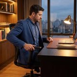 The image features an ergonomic office chair designed for comfort and support during long hours of work. A man in a blue blazer is seated at a desk, using a laptop, with a modern office setting visible in the background. The chair's design includes adjustable armrests, emphasizing its ergonomic features.