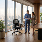 The image features an ergonomic office chair, designed for comfort and support during long hours of work. The chair has a mesh back for breathability and is positioned in a modern office space with large windows showcasing a city skyline. Two professionals are engaged in discussion, holding documents and a tablet, suggesting a collaborative work environment.