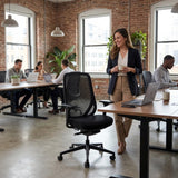 The product is an ergonomic office chair, likely from the "Glory" model line. The image shows a modern office environment with a woman standing and holding a cup, while several people are seated at desks working on laptops. The chair features a mesh back and a black seat, emphasizing comfort and support in a professional setting.