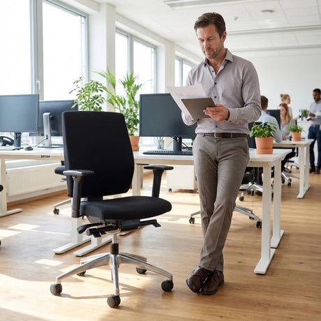 The image features an ergonomic office chair, specifically the "Profi" model. It showcases a sleek black design with adjustable armrests and a five-star base on wheels. In the background, a modern office setting is visible, with several desks, computers, and plants, creating a professional atmosphere. A man is standing beside the chair, reviewing documents.