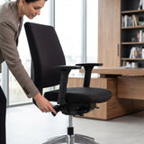 The image features an ergonomic office chair, likely designed for comfort and support during long hours of sitting. A woman is adjusting the chair's height or tilt using a lever, showcasing its ergonomic features. The setting appears to be a modern office with wooden furniture and large windows, emphasizing a professional environment.