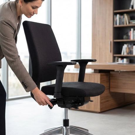 The image features an ergonomic office chair, likely designed for comfort and support during long hours of sitting. A woman is adjusting the chair's height or tilt using a lever, showcasing its ergonomic features. The setting appears to be a modern office with wooden furniture and large windows, emphasizing a professional environment.