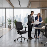 The image features an ergonomic office chair, likely from a professional brand, designed for comfort and support during long hours of work. In the scene, two individuals are discussing plans while standing next to a modern office setup, which includes the chair prominently displayed. The backdrop showcases a city skyline through large windows, enhancing the contemporary office atmosphere.
