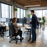 The image features an ergonomic office chair, likely from the brand Rapthor. In the scene, a woman is seated at a desk with multiple computer monitors, engaged in conversation with a man standing nearby. The office environment is modern, with large windows and plants, creating a bright and inviting workspace.