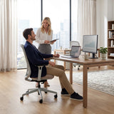 The image features an ergonomic office chair, designed for comfort and support during long hours of work. A man is seated in the chair at a modern desk, while a woman stands nearby, holding a tablet. The workspace is bright and contemporary, with large windows showcasing a city view, and there are two computers on the desk displaying data graphs.