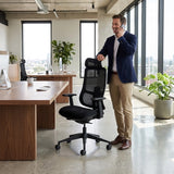 The product is an ergonomic office chair, specifically the Valor model. The image shows a man standing next to the chair in a modern office setting, engaged in a phone call. The chair features a mesh back for breathability and adjustable armrests, positioned in front of a wooden desk with a computer and plants in the background.