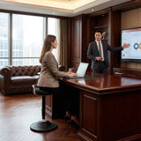 The image features a sit-stand stool designed for ergonomic office use. It appears to be a balance stool that allows for dynamic sitting and promotes better posture. In the scene, a woman is seated on the stool while working on a laptop at a wooden desk, and a man is presenting information on a screen in the background. The office setting is modern
