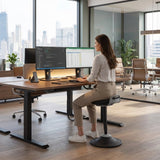 The image features a sit-stand stool designed for ergonomic office use. It appears to be a balance stool that allows for dynamic sitting, promoting better posture while working. A woman is seen using the stool at a modern desk with dual monitors, showcasing a contemporary office environment with large windows and city views in the background.