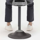 The image depicts a sit-stand stool designed for ergonomic support, promoting active sitting. The stool features a sleek design with a circular base and an adjustable height mechanism. In the image, a person is shown using the stool, wearing dark pants and white shoes, emphasizing the product's use in a modern workspace.