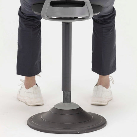 The image depicts a sit-stand stool designed for ergonomic support, promoting active sitting. The stool features a sleek design with a circular base and an adjustable height mechanism. In the image, a person is shown using the stool, wearing dark pants and white shoes, emphasizing the product's use in a modern workspace.