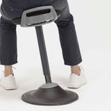 The product is a sit-stand stool designed for ergonomic support, allowing users to alternate between sitting and standing positions. The image shows a person using the stool, which features a sleek design with a padded seat and a sturdy base, promoting balance and comfort during use.