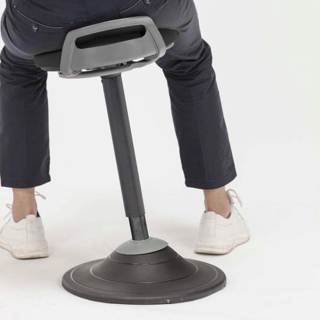 The product is a sit-stand stool designed for ergonomic support, allowing users to alternate between sitting and standing positions. The image shows a person using the stool, which features a sleek design with a padded seat and a sturdy base, promoting balance and comfort during use.