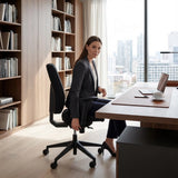The image features an ergonomic office chair, designed for comfort and support during long hours of work. A woman is seated in the chair, which has adjustable features, while positioned at a modern desk with a laptop and a cup of coffee. The background shows a stylish office space with bookshelves and a cityscape view.