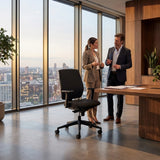 The image features an ergonomic office chair, specifically the "ergonomic-office-chair-basic" model. The chair has a sleek design with a black fabric covering and is positioned in a modern office setting. Two professionals, a man and a woman, are engaged in conversation near a large window that offers a city view, highlighting a contemporary and stylish workspace.