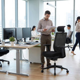 The image features an ergonomic office chair designed for comfort and support during long working hours. The chair is black with a mesh backrest, promoting breathability. In the scene, a man is standing and reviewing documents while two other individuals work at desks with multiple monitors in a modern office setting. The workspace is bright and open, with plants adding a touch