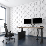 The image features an ergonomic office chair, designed for comfort and support during long hours of work. The chair is positioned next to a modern standing desk equipped with dual monitors. The room has a sleek, contemporary aesthetic with a textured white wall and large windows allowing natural light to enter.