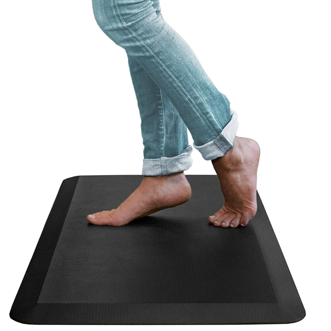 The image shows an ergonomic anti-fatigue mat designed for standing workspaces. It features a textured black surface and is positioned on the floor, with a person's feet partially visible as they stand on it. The mat aims to provide comfort and support to reduce fatigue during prolonged standing. The brand and model are not specified in the image.