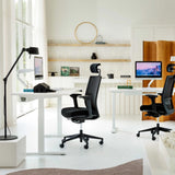 The image features an ergonomic office chair, likely designed for comfort and support during long hours of work. The brand and model are not specified, but the chair is black with a high back and adjustable features. In the background, there are two desks with monitors, a stylish lamp, and decorative items on shelves, creating a modern office environment.