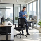 The image features an ergonomic office chair, specifically the UVI Energetic model. It showcases a modern office environment with a man standing beside the chair while reviewing documents. The chair is black with a mesh back, and the workspace includes desks with computers and plants, creating a professional and inviting atmosphere.