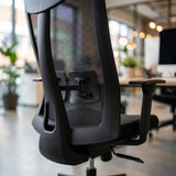 The product is an ergonomic office chair, specifically the UVI Energetic model. The image shows the back of the chair, highlighting its mesh design and adjustable features, including armrests and a lever for height adjustment. The setting appears to be a modern office space with plants and soft lighting in the background.