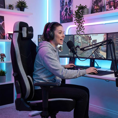The image features a gaming ergonomic office chair from the brand UVI, specifically the UVI Alpha model. A woman is seated in the chair, wearing a hoodie and headphones, engaged in gaming at a dual-monitor setup. The background showcases a modern gaming environment with colorful LED lighting and decorative items.