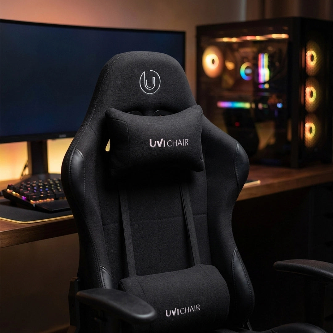 The product is an ergonomic office chair designed for gaming, specifically the UviChair model in black. The image showcases the chair's sleek design, featuring a high backrest, a supportive neck pillow, and a modern aesthetic. In the background, there is a gaming setup with a keyboard and a colorful PC case, enhancing the gaming atmosphere.