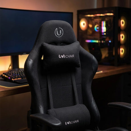 The product is an ergonomic office chair designed for gaming, specifically the UviChair model in black. The image showcases the chair's sleek design, featuring a high backrest, a supportive neck pillow, and a modern aesthetic. In the background, there is a gaming setup with a keyboard and a colorful PC case, enhancing the gaming atmosphere.