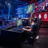 The image features a gaming ergonomic office chair, specifically the UVI model in black. It shows two gamers in a competitive esports setting, with one player focused on a monitor displaying a game, while another points at the screen. The environment is lively, with a large audience in the background and vibrant stage lighting.