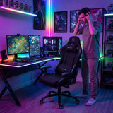 The image features a gaming ergonomic office chair, specifically the UVI chair model in black. The scene includes a person standing near the chair, which is positioned in a colorful gaming setup with dual monitors displaying a game, a gaming PC with RGB lighting, and ambient lighting that enhances the gaming atmosphere.