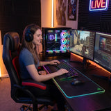 The image features an ergonomic gaming office chair, specifically the UVI Hero model in red. A woman is sitting comfortably in the chair while using a dual-monitor setup for gaming, complete with a colorful gaming PC and RGB lighting in the background. She is wearing headphones and appears engaged in her gaming experience.