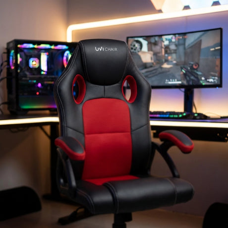The product is an ergonomic office chair designed for gaming, specifically the UVI HERO model in red and black. The chair features a sporty design with cut-out holes in the backrest for ventilation, and it is positioned in a gaming setup with a colorful computer setup and a monitor displaying a game.