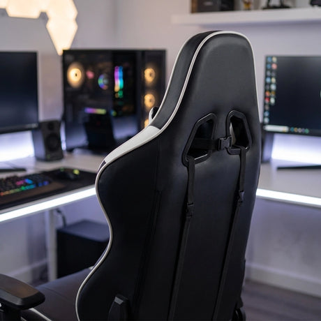 The image features a gaming chair, specifically the UVI Sport XL model in white and black. The chair has an ergonomic design with a high backrest and is positioned in a modern gaming setup, showcasing multiple monitors and RGB lighting in the background.
